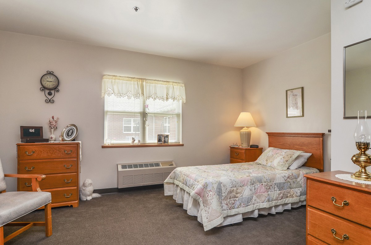 A cozy bedroom with a neatly made bed, dresser, and a lamp beside the window.