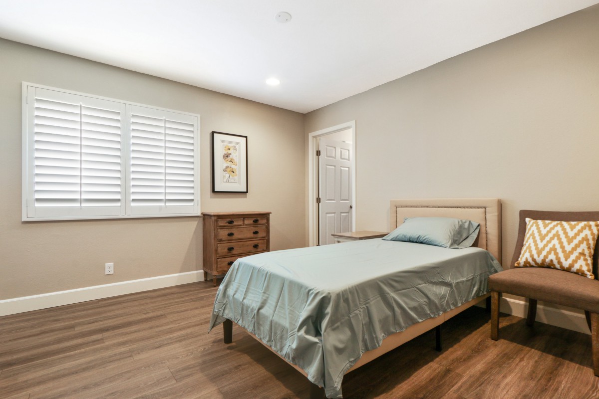 A minimalist bedroom featuring a single bed, wooden floors, and a small dresser under a window with shutters.