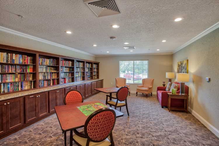 A cozy library room with bookshelves, tables, and chairs, featuring a large window providing natural light.