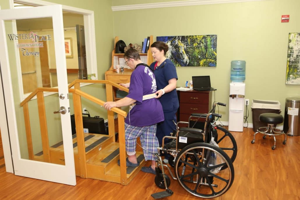 A caregiver assists a person in a physical therapy session with a wheelchair nearby.