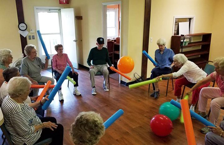 A group of seniors engaging in a playful indoor exercise using foam noodles and therapy balls.