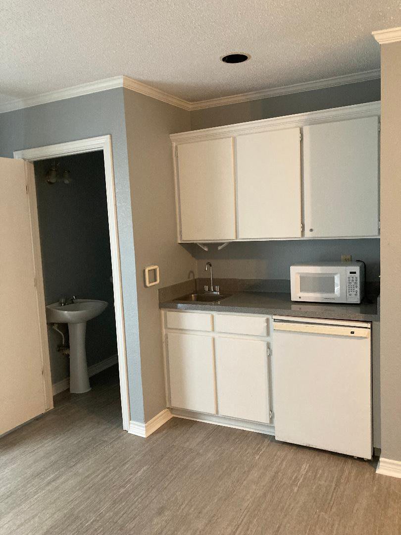 A compact kitchenette with white cabinets, a microwave, and a small refrigerator adjacent to an open bathroom door.