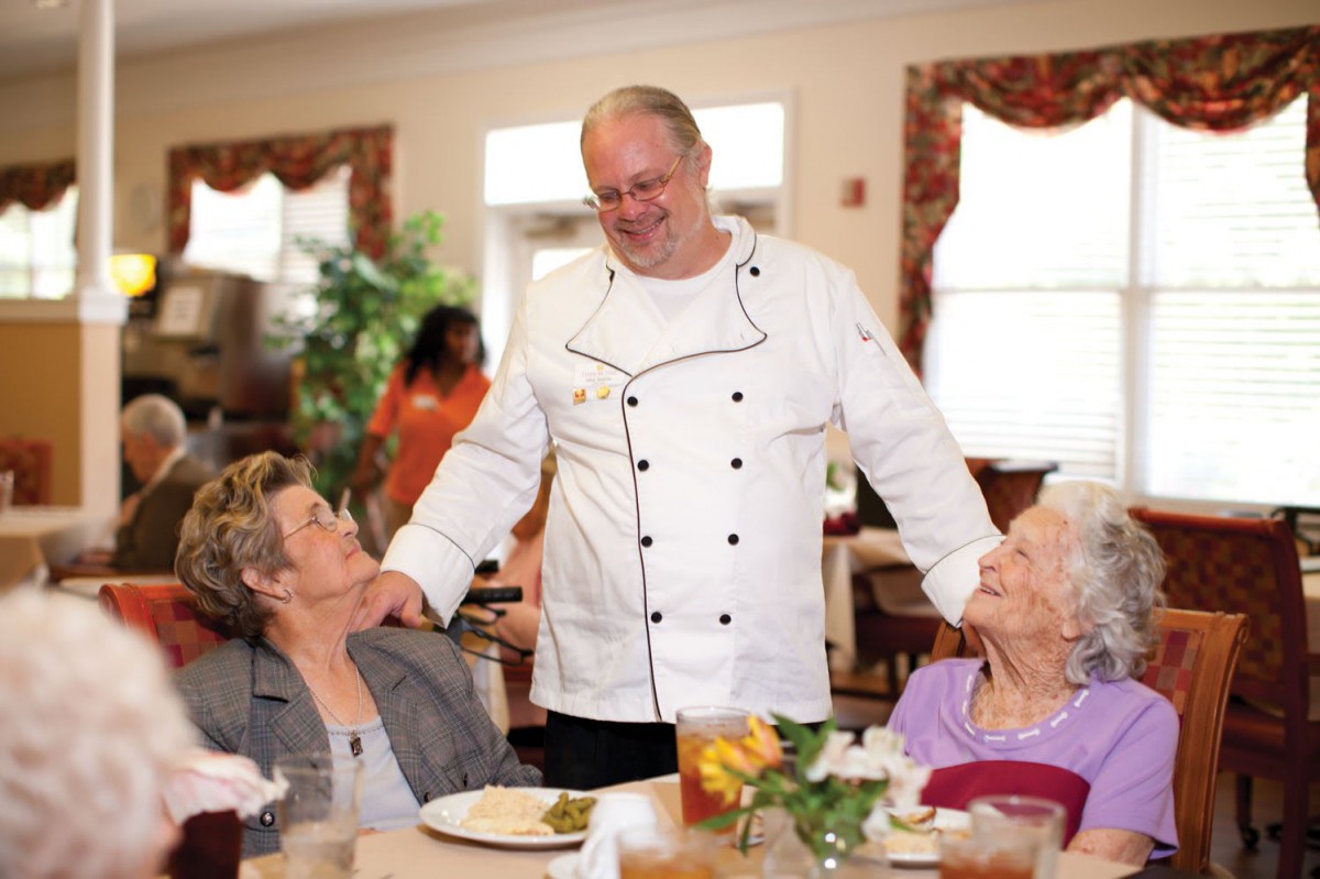 A chef interacts with two elderly women in a dining setting, showcasing hospitality.