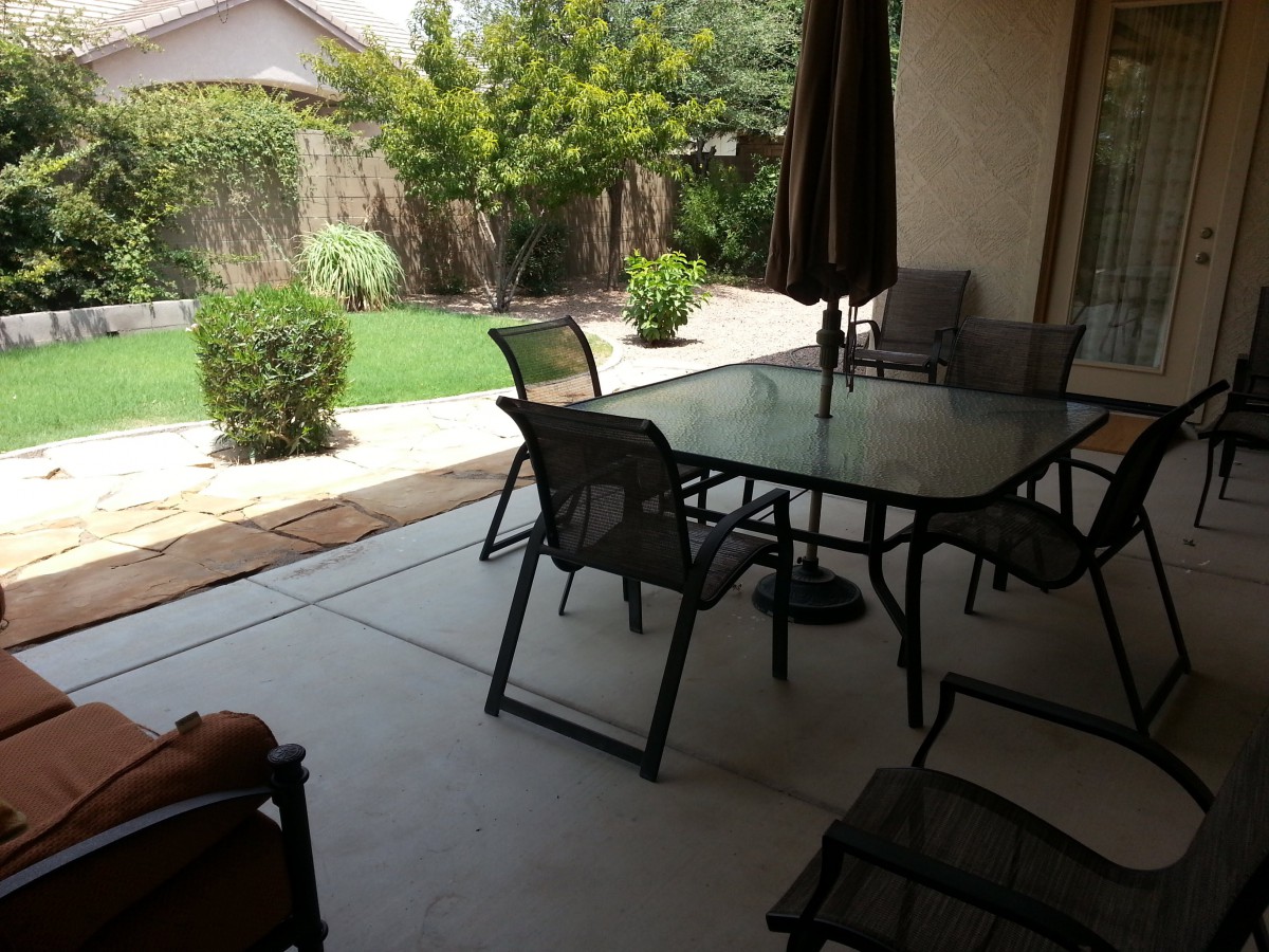 A cozy backyard patio featuring a glass table with chairs and an umbrella, overlooking a lush garden.