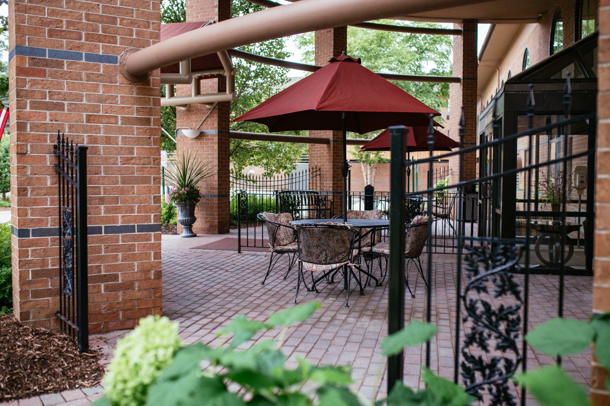A cozy patio with tables, chairs, and a red umbrella set against a backdrop of brickwork and greenery.