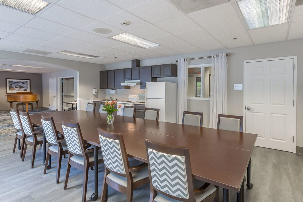 A modern conference room with a long meeting table and chairs, adjacent to a kitchen area.