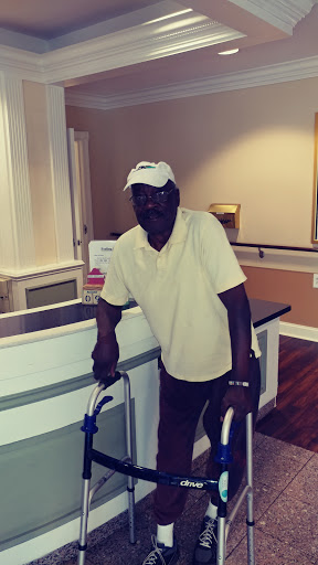 An older man using a walking aid stands by a reception area, dressed casually in a yellow shirt and white cap.