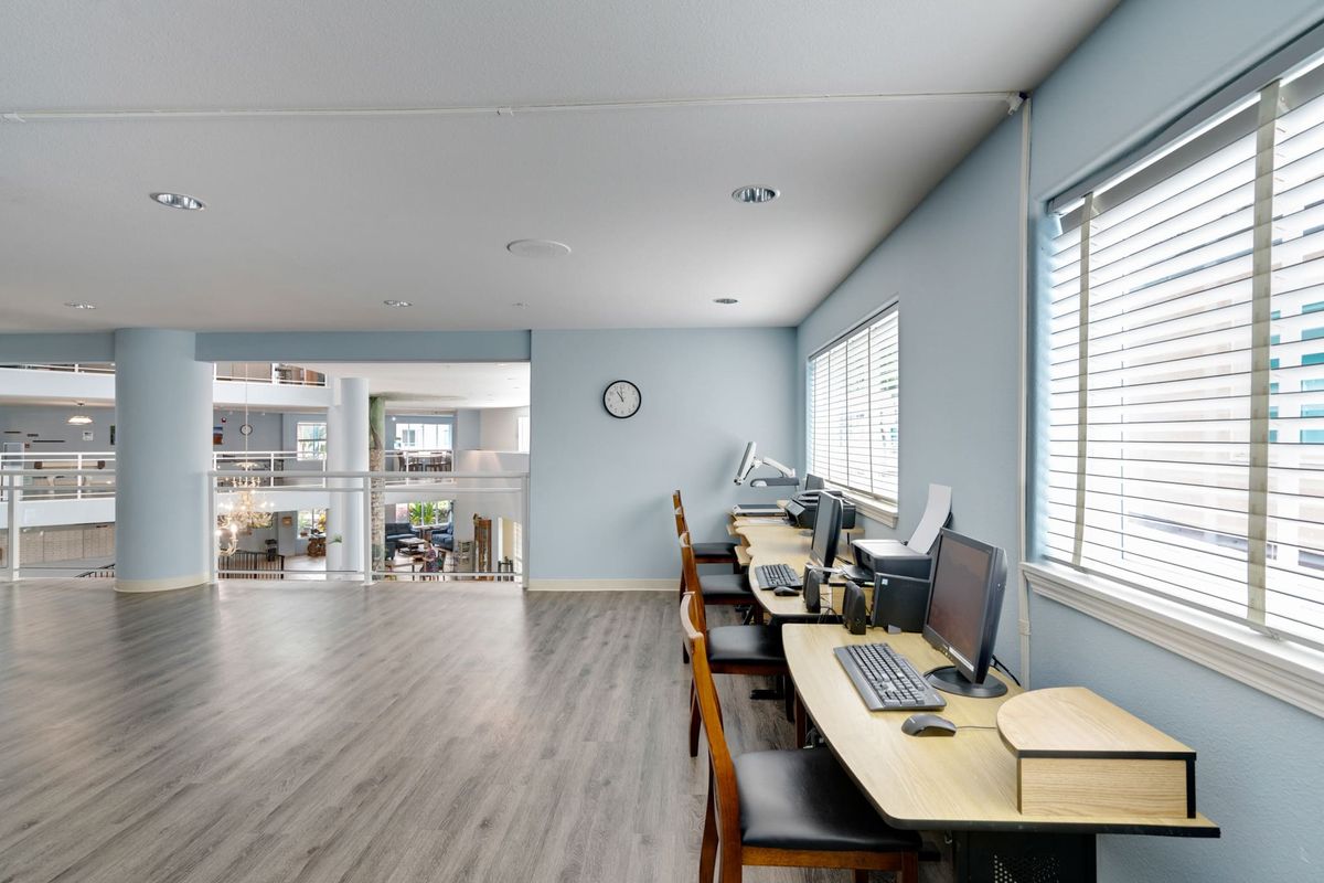A modern office workspace with desks, computers, and chairs along a windowed wall.