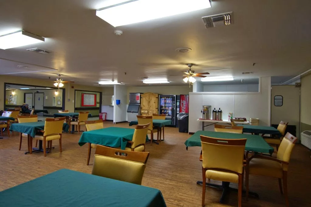 A spacious dining area with green tablecloths and yellow chairs, featuring a beverage station in the background.