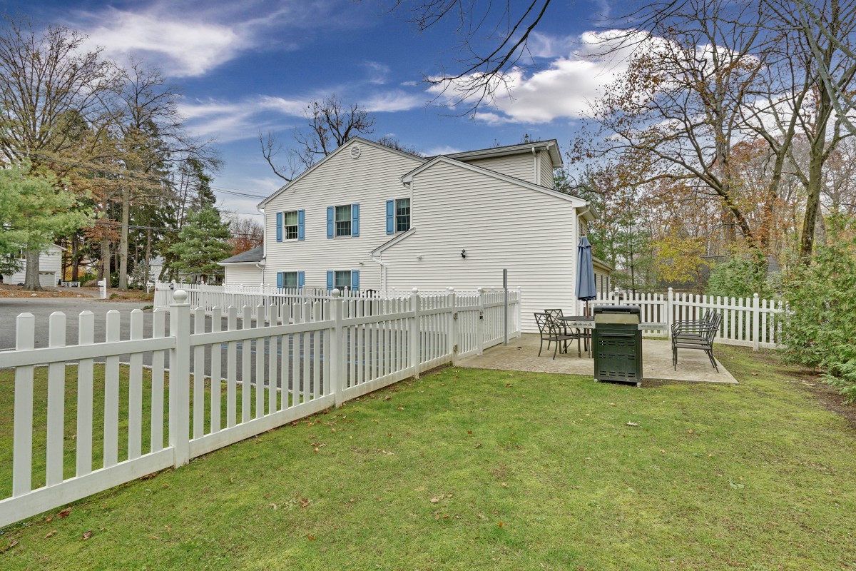 A white house with blue shutters, surrounded by a fenced yard with a patio area.