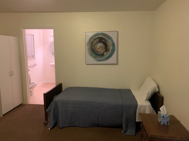 A simple bedroom with a single bed, artwork on the wall, and an open door leading to an adjacent room.