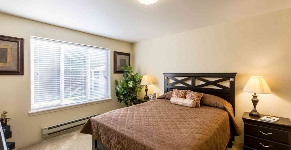 A cozy bedroom with a large window, featuring a bed with a brown quilt and pillows, complemented by a lamp and a potted plant.