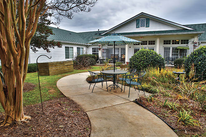A cozy outdoor patio area with a table and umbrella, surrounded by greenery and a pathway leading to the house.