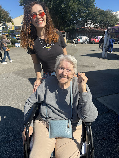 A young woman joyfully poses with an elderly lady in a wheelchair on a sunny day.