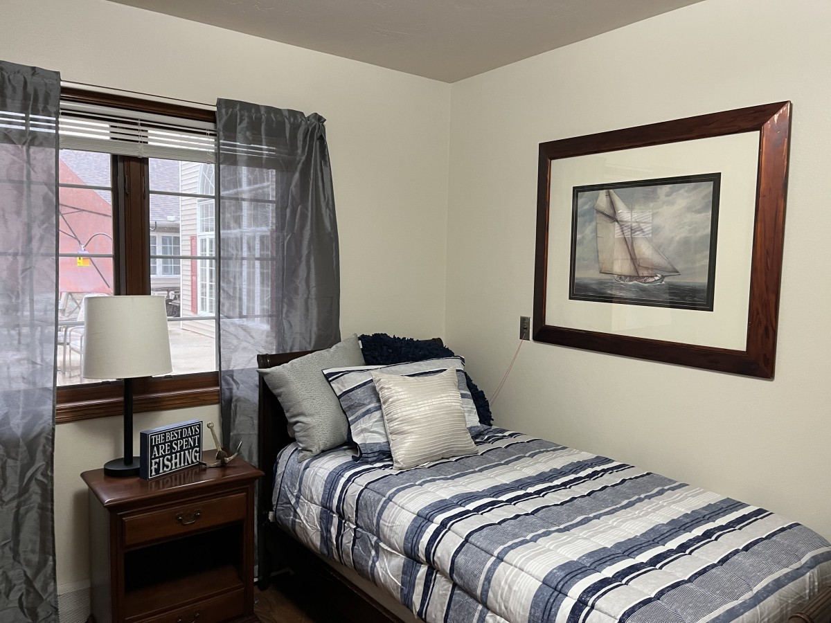 A cozy bedroom with a neatly made single bed, featuring striped bedding and decorative pillows. A nightstand with a lamp sits beside the bed, and a large painting hangs on the wall.