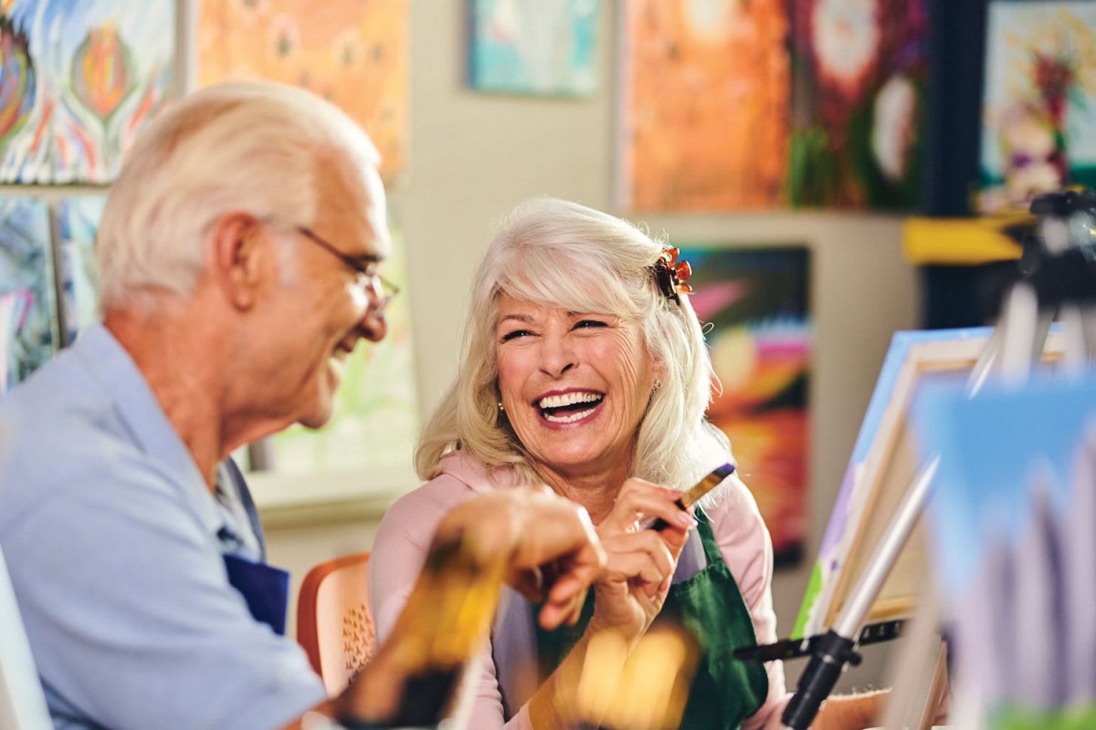 An elderly couple laughing together while painting in an art class.
