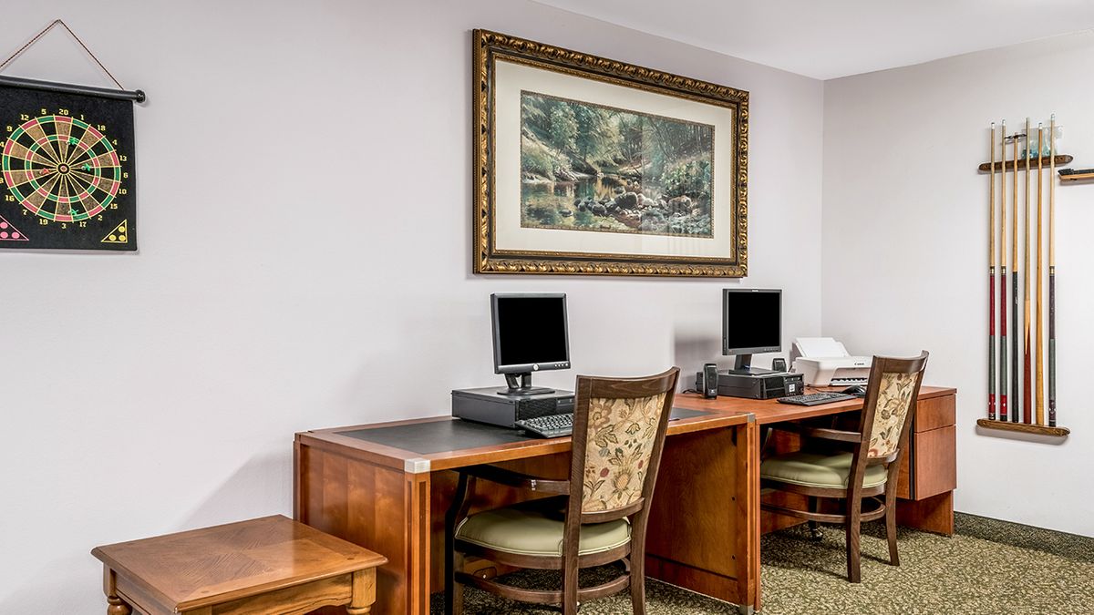 An office space with computers on wooden desks and chairs, featuring framed artwork on the wall.