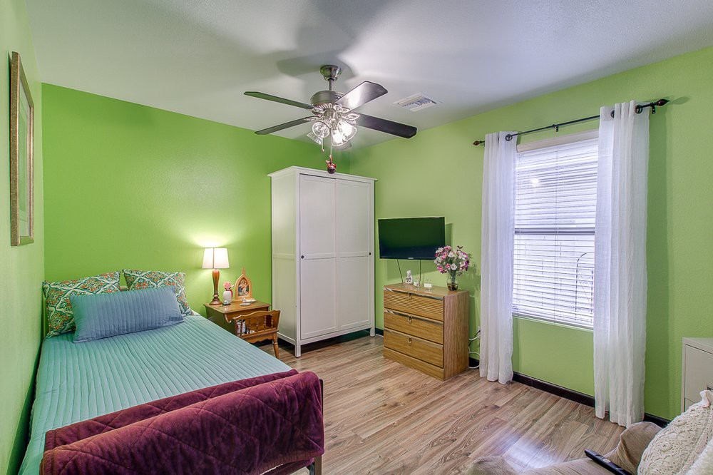 A cozy bedroom with vibrant green walls, featuring a bed, a wardrobe, and a dresser, illuminated by natural light through a window.