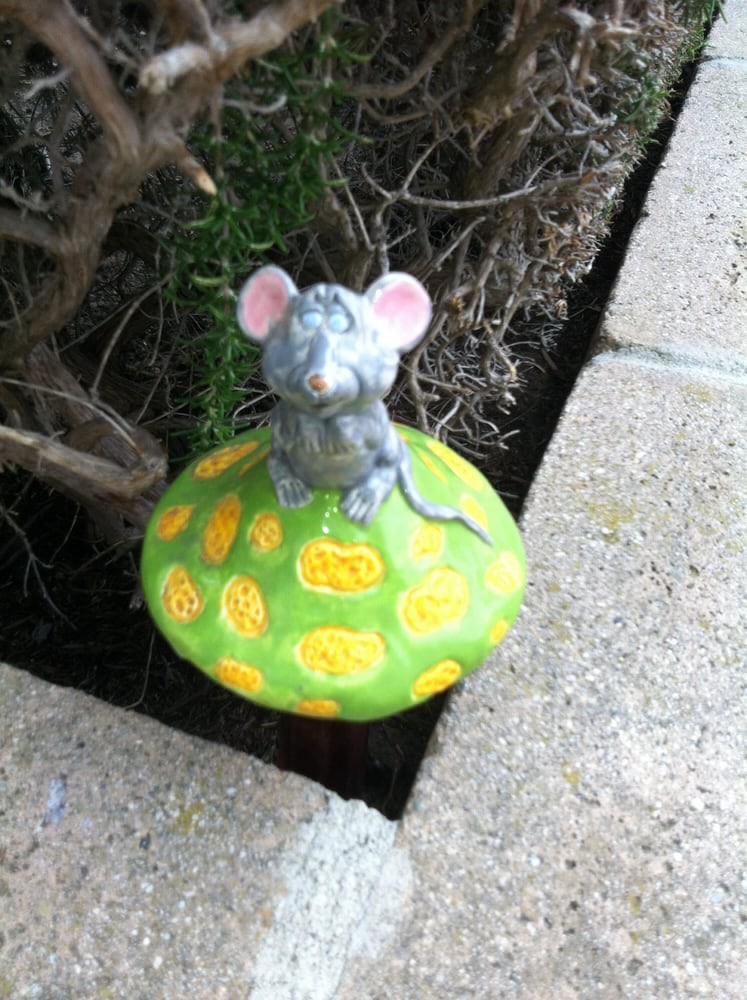 A mouse figurine sitting on a colorful mushroom in a garden setting.