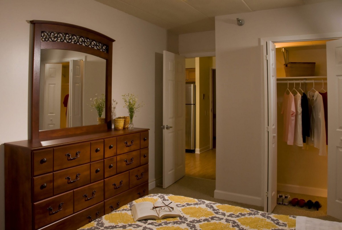A cozy bedroom with a wooden dresser and a mirror, featuring a patterned bedspread and open doorways.