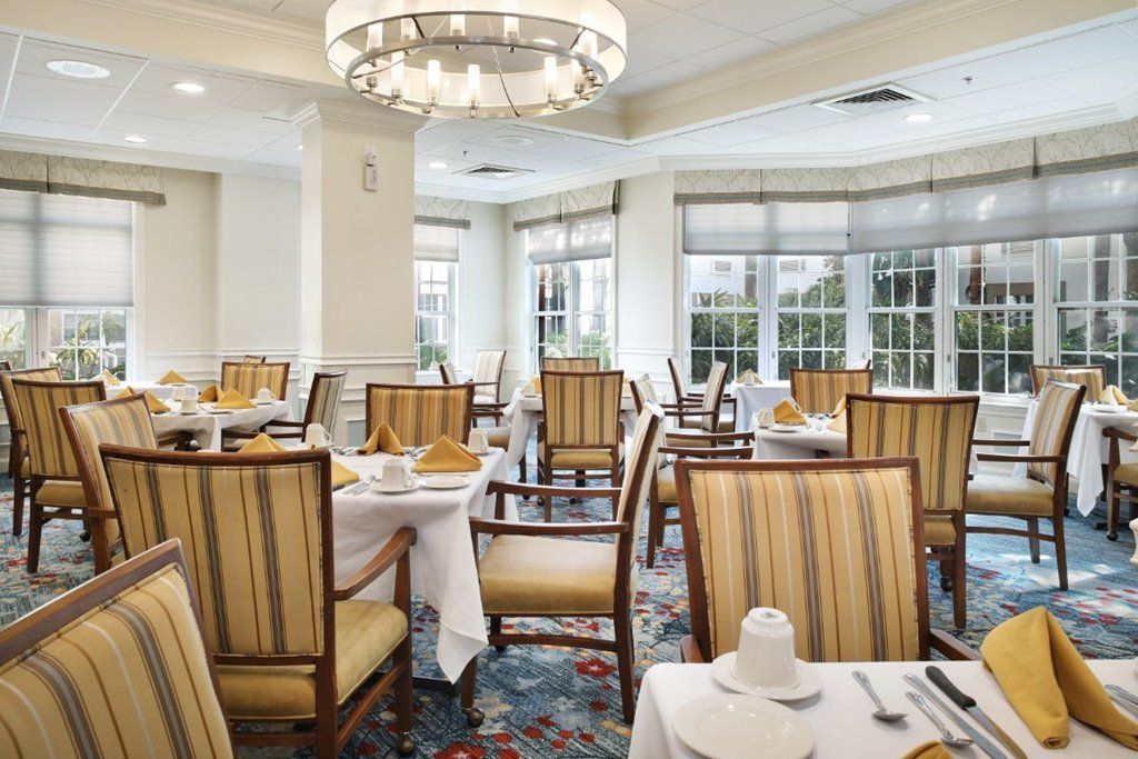 A well-lit dining room with neatly arranged tables and chairs, featuring large windows and a decorative chandelier.