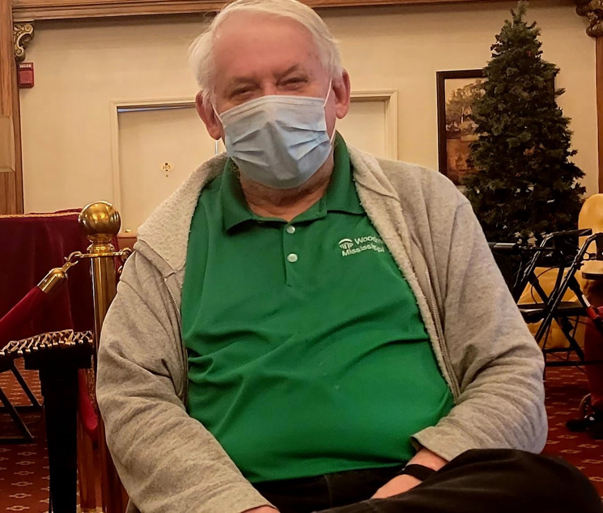 A man wearing a mask, seated indoors in casual attire.