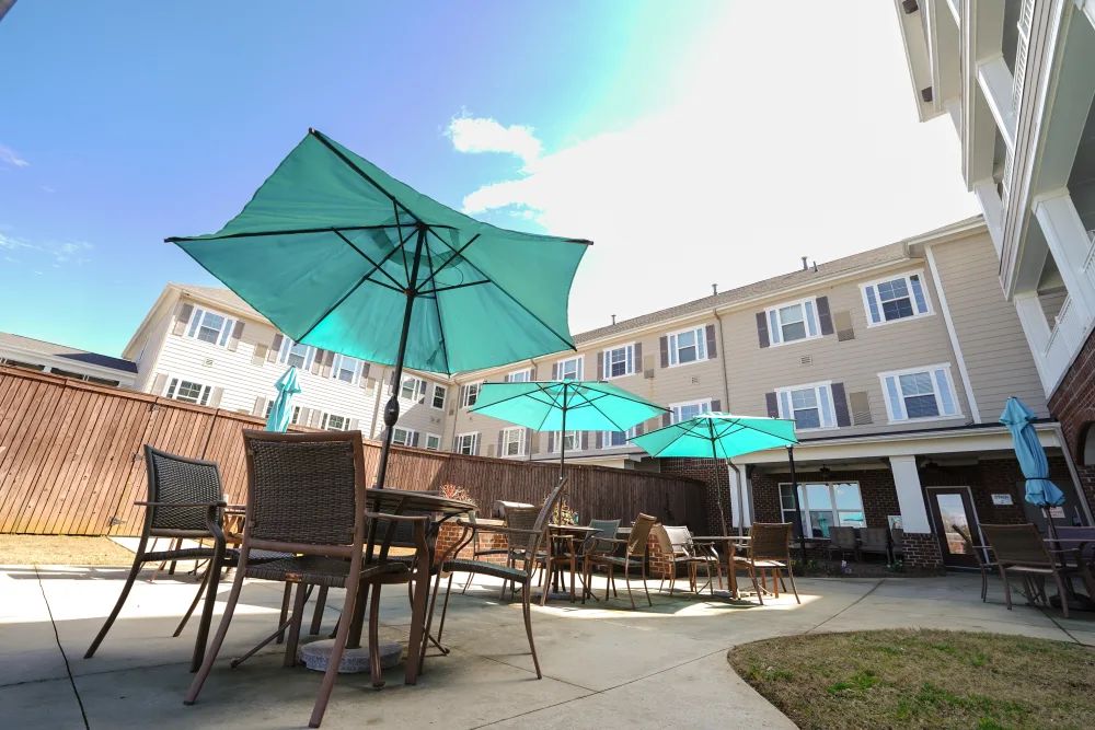 A sunny patio area with tables, chairs, and teal umbrellas, adjacent to a multi-story building.