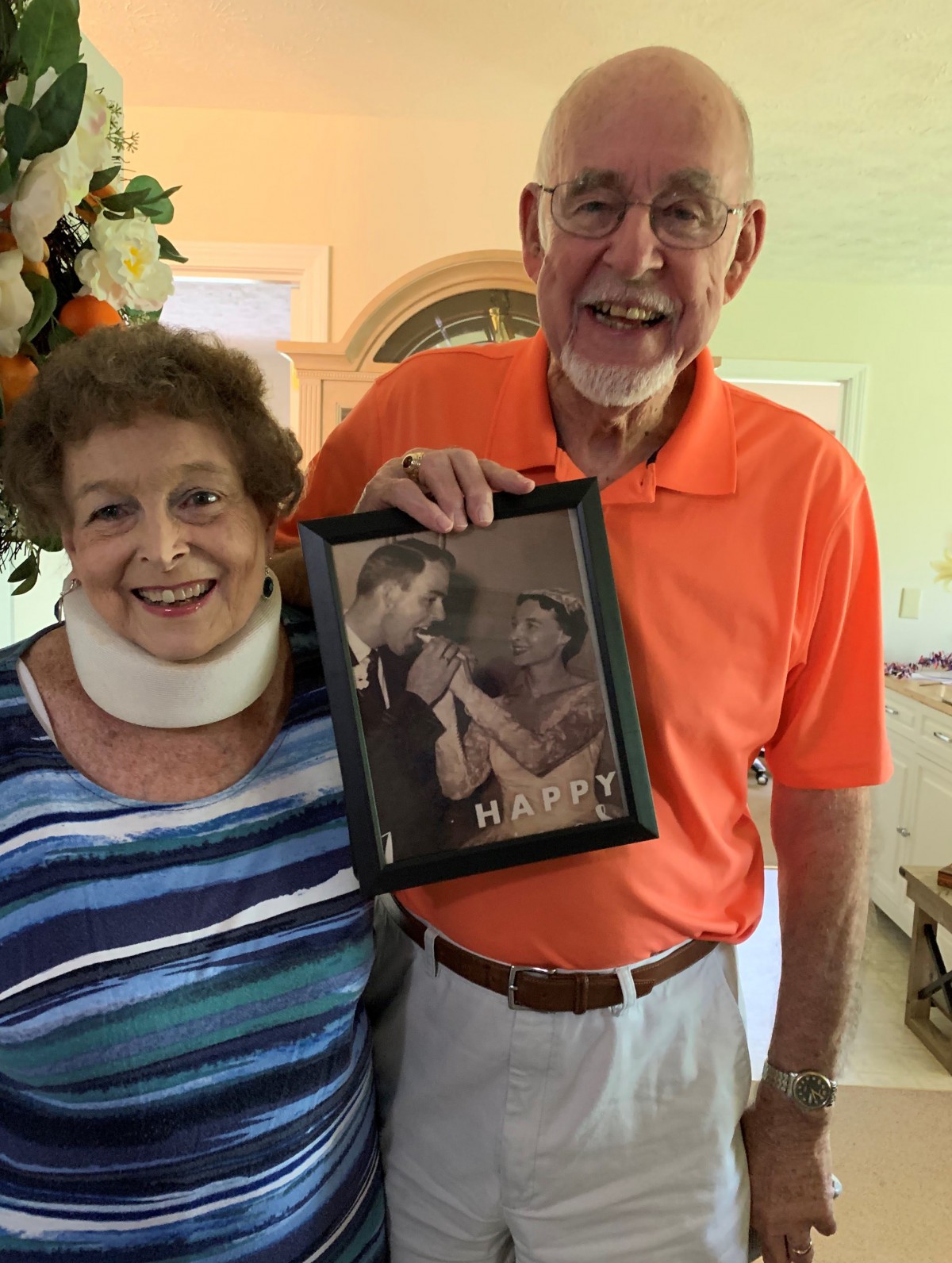An elderly couple smiles while holding a framed photograph of themselves.