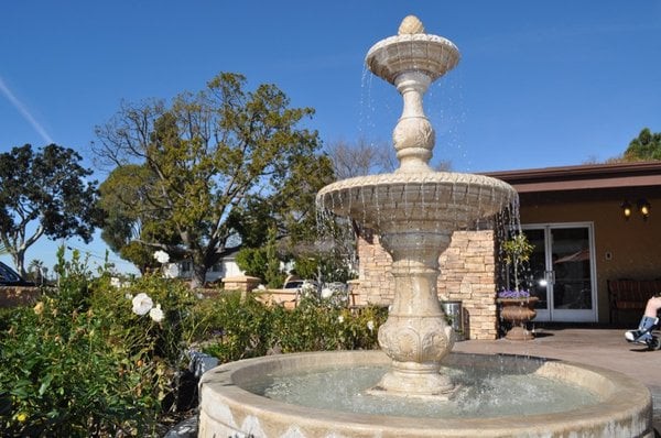 A stone fountain in a garden setting, surrounded by greenery and flowers, under a clear blue sky.