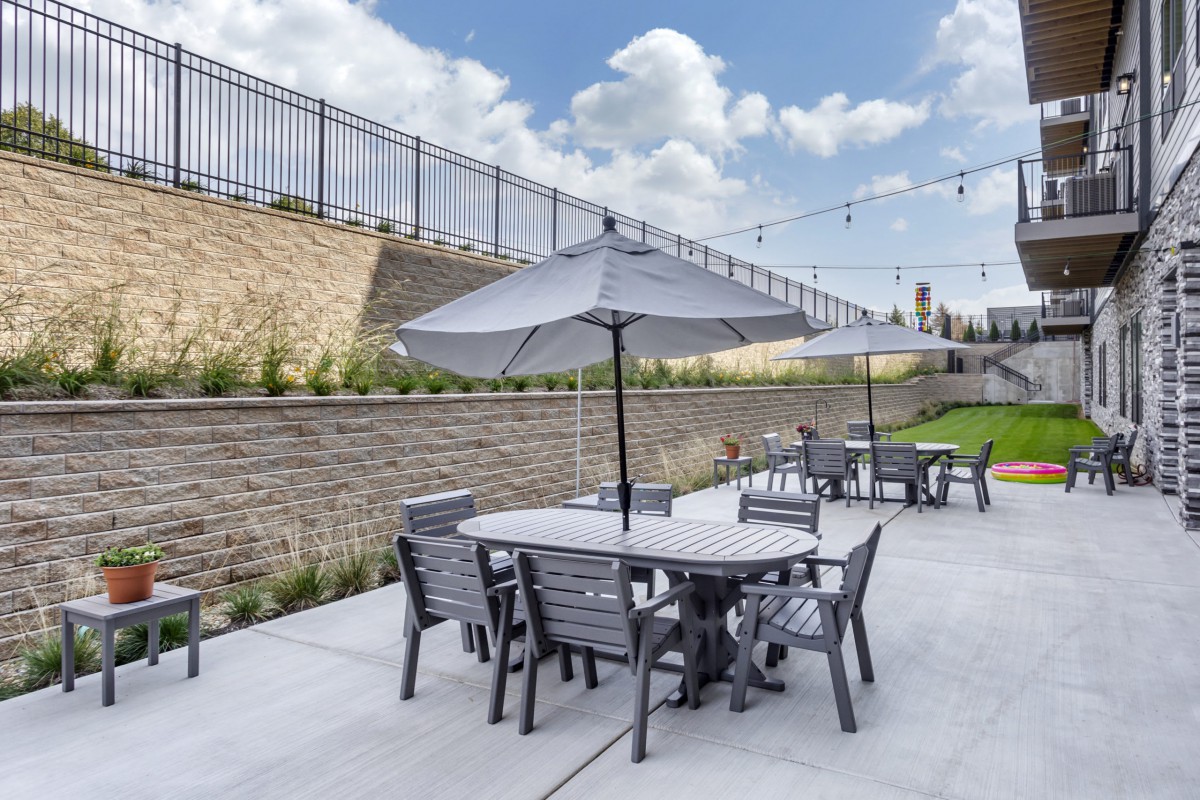 A modern outdoor patio with tables and chairs under umbrellas, surrounded by a garden and string lights.