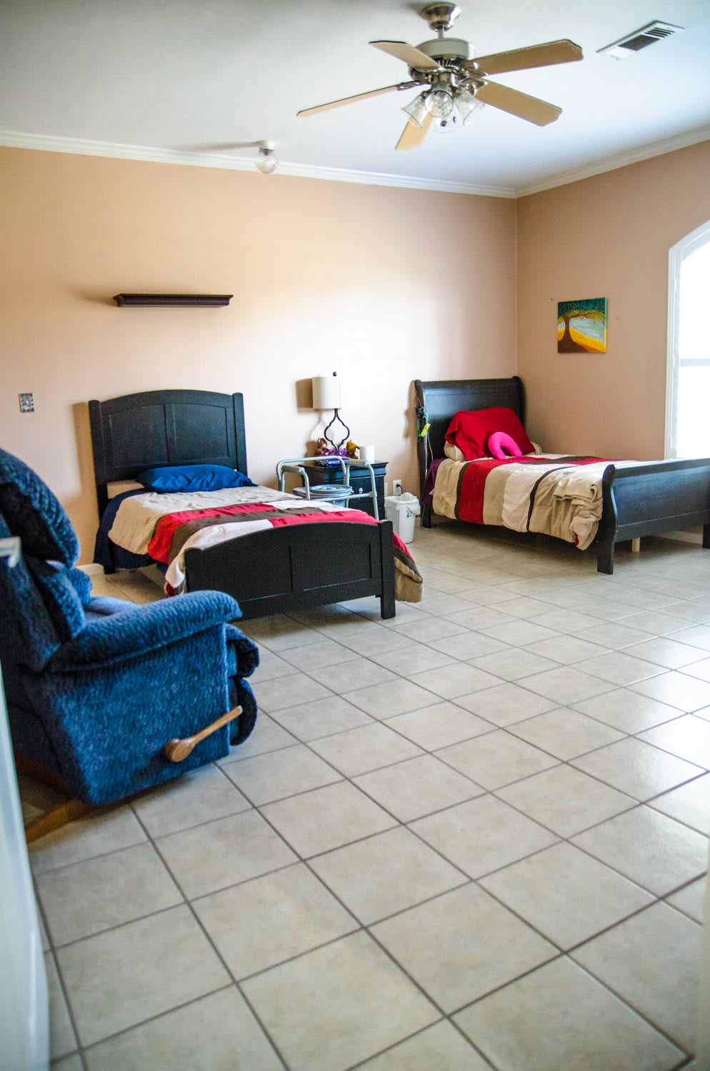 A spacious bedroom with two beds, a blue armchair, and tiled flooring.