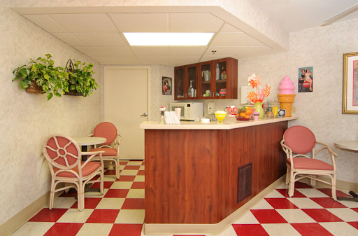 A cozy retro kitchen area featuring a wooden bar, pink chairs, and a vibrant checkered floor.