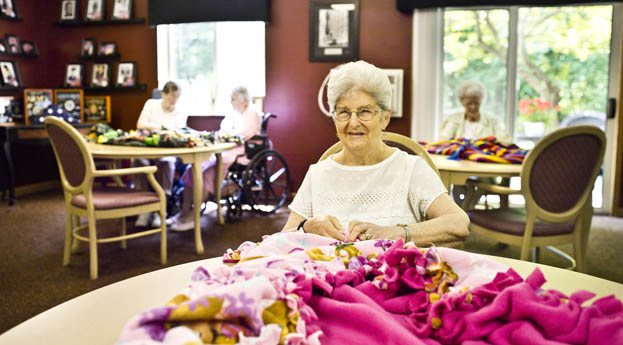 An elderly woman is smiling while engaged in a crafting activity at a community center.