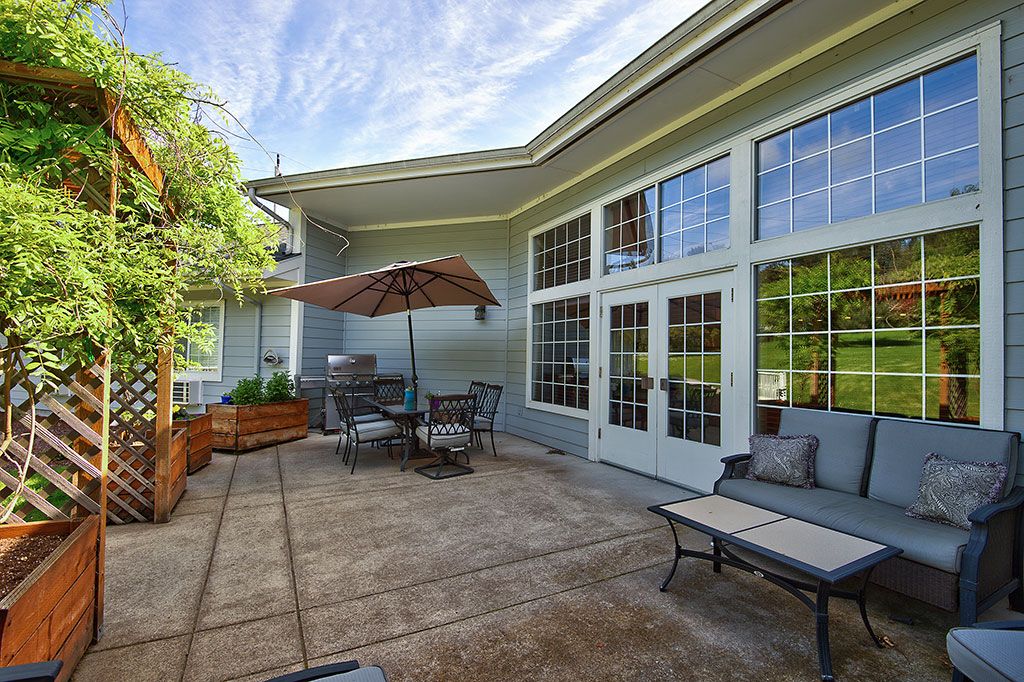 A cozy outdoor patio with seating and an umbrella, surrounded by greenery and large windows.