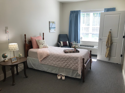 A cozy and well-lit bedroom featuring a single bed with soft pink bedding, a nightstand, and a comfortable chair.