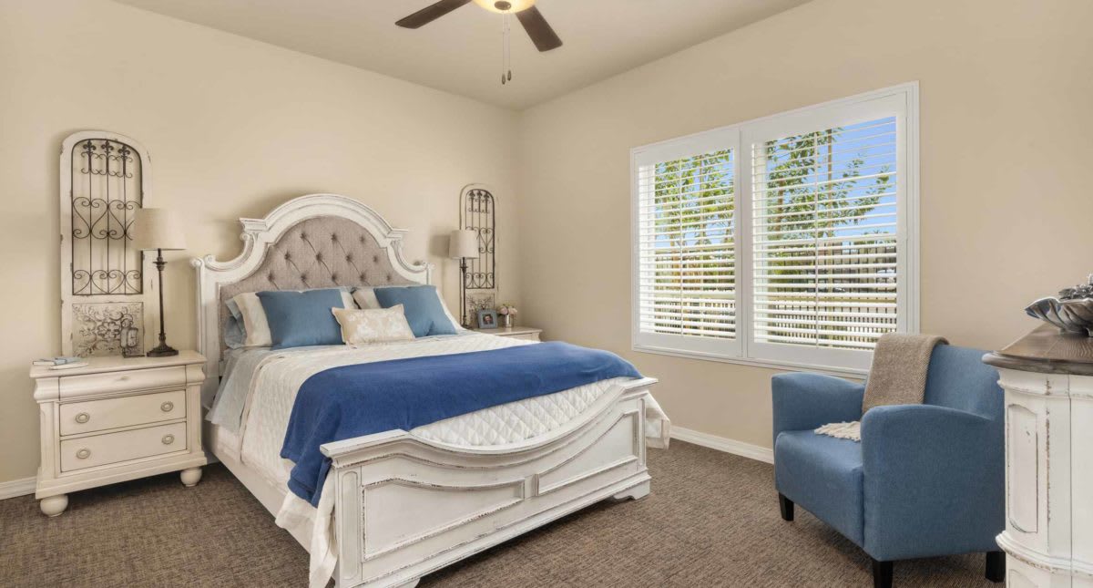 A cozy bedroom featuring a white bed with blue bedding, an armchair, and large windows with blinds.