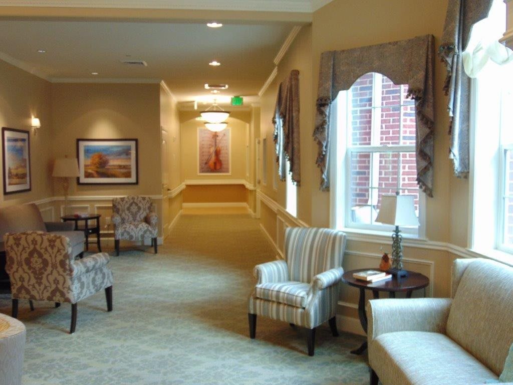 A warmly lit hallway featuring comfortable chairs, large windows with drapes, and decorative paintings on the walls.