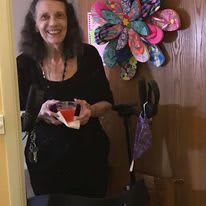A woman smiling indoors, holding a drink with colorful flower decoration on the wall.