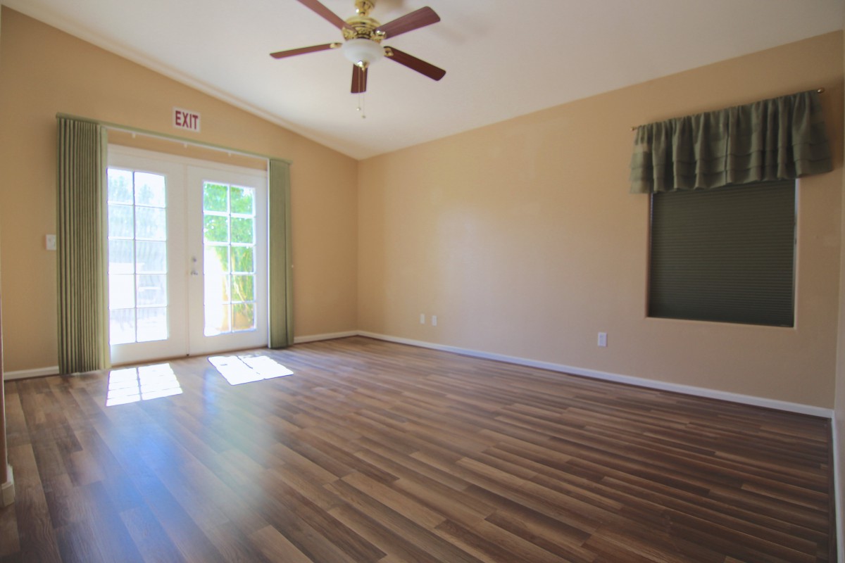 A spacious empty room with hardwood flooring and a ceiling fan, featuring large windows allowing natural light.