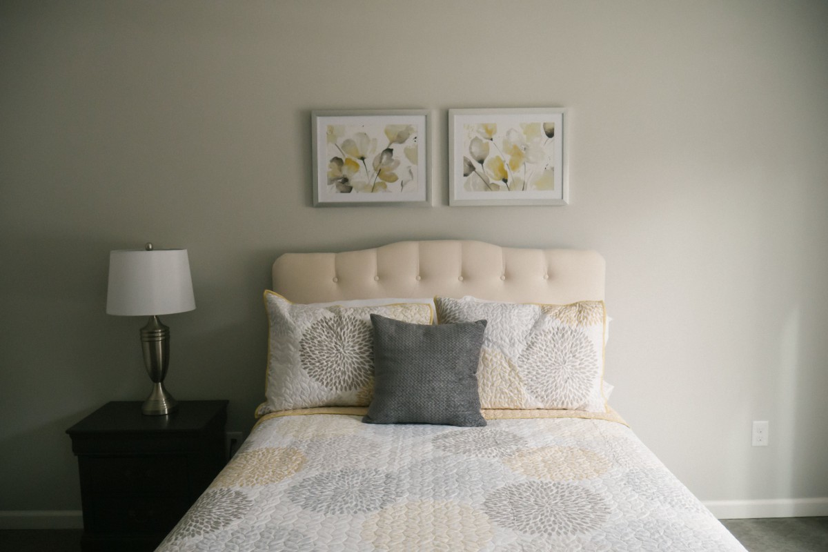 A neatly arranged bedroom with a cushioned headboard, decorative pillows, and framed artwork above the bed.