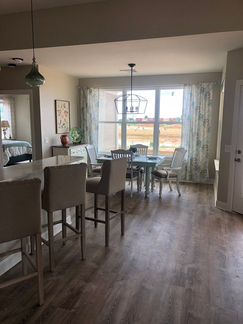 A bright and airy dining area connected to the kitchen, featuring a large window with curtains, a dining table with chairs, and a bar counter with stools on a hardwood floor.