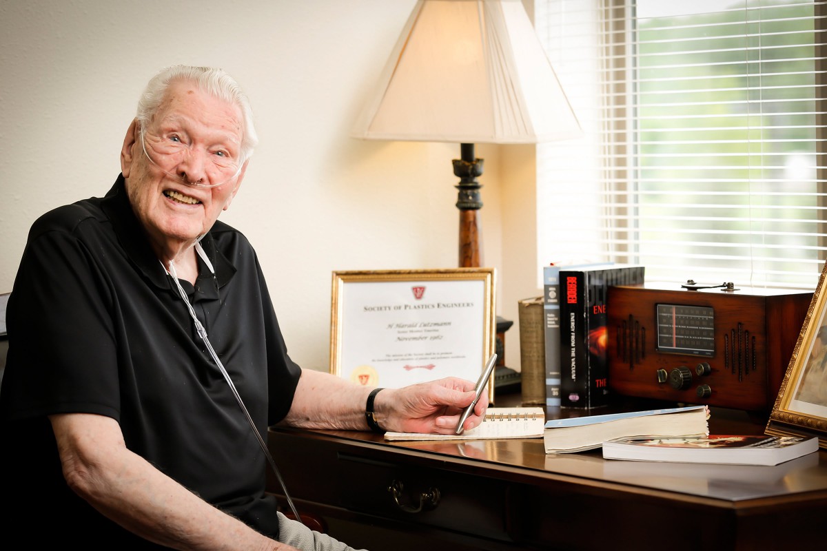 An elderly man smiles while sitting at a desk with books and a certificate nearby.