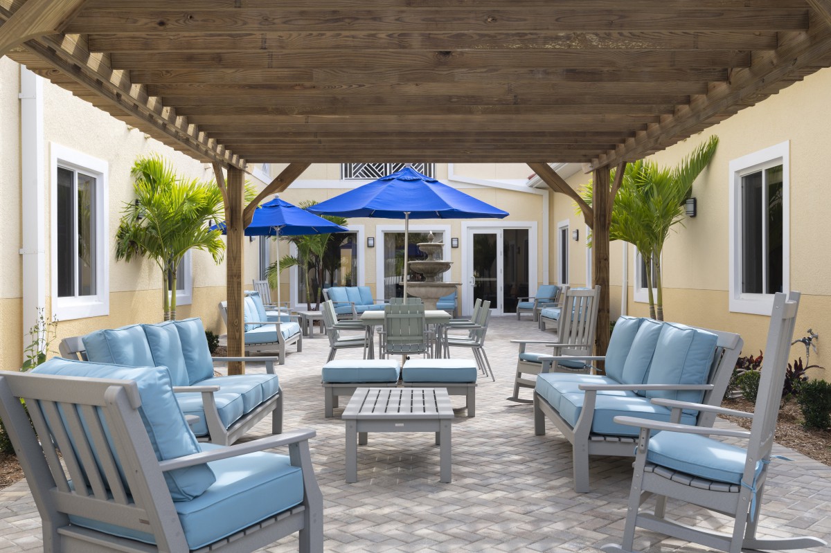 A cozy outdoor patio with cushioned seating under a wooden pergola and blue umbrellas, surrounded by palm trees.