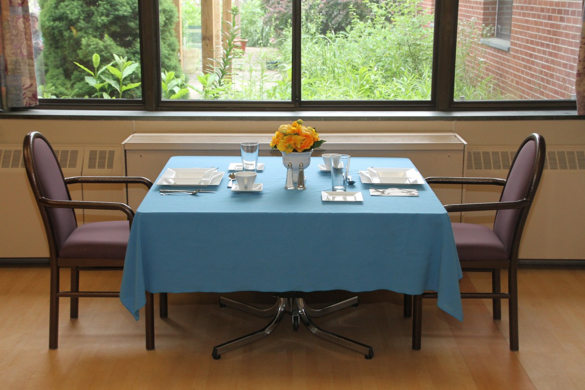 A dining table set with a blue tablecloth, complete with cutlery, glasses, and a flower arrangement, near a large window overlooking greenery.