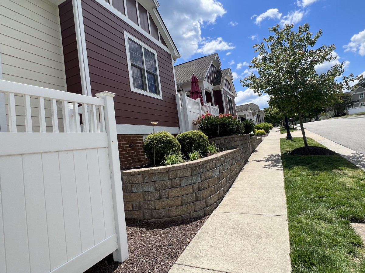 A sunny suburban street with well-maintained houses, landscaping, and a white fence.
