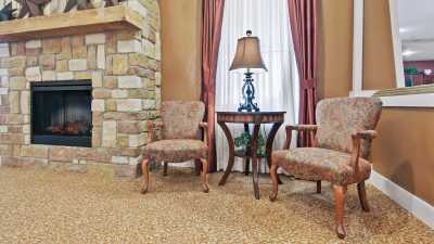 A cozy living room area with two antique-style chairs flanking a small round table and lamp, complemented by a stone fireplace and elegant curtains.