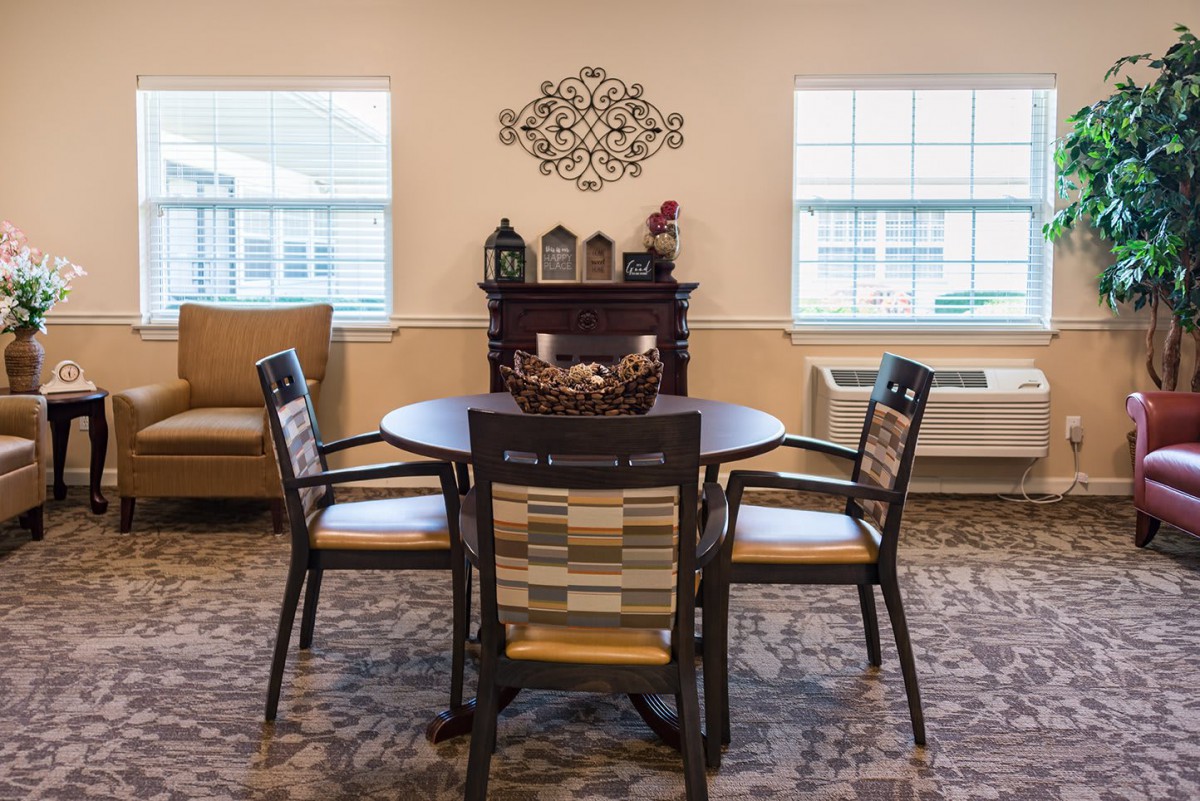 A cozy dining area with a round table and four chairs, decorated with wall art and lit by natural light from two windows.