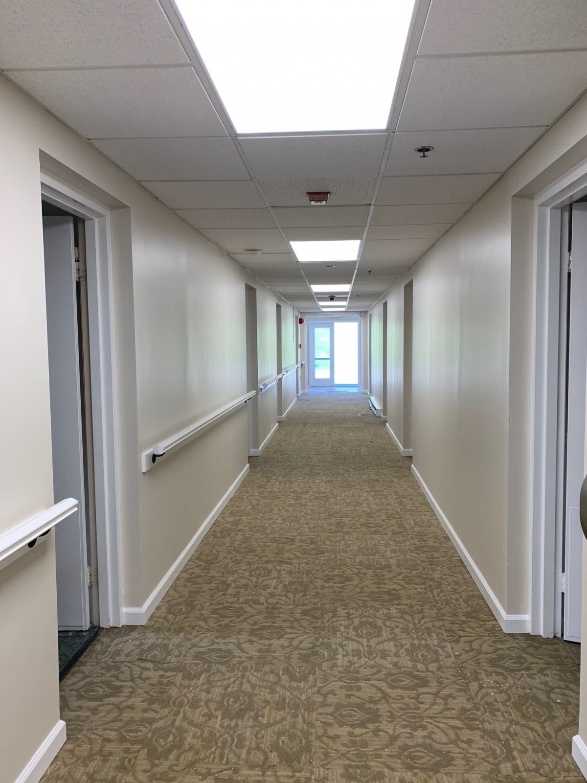 A long indoor hallway with carpet, doors on both sides, and overhead lighting.
