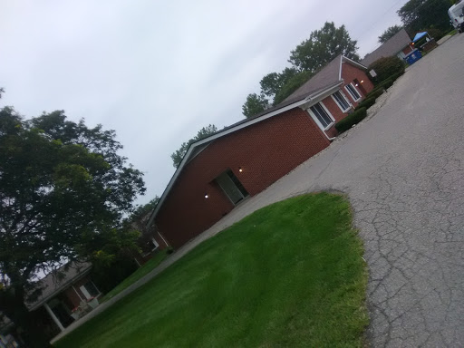 A peaceful residential area featuring a brick house surrounded by green grass and trees under a cloudy sky.
