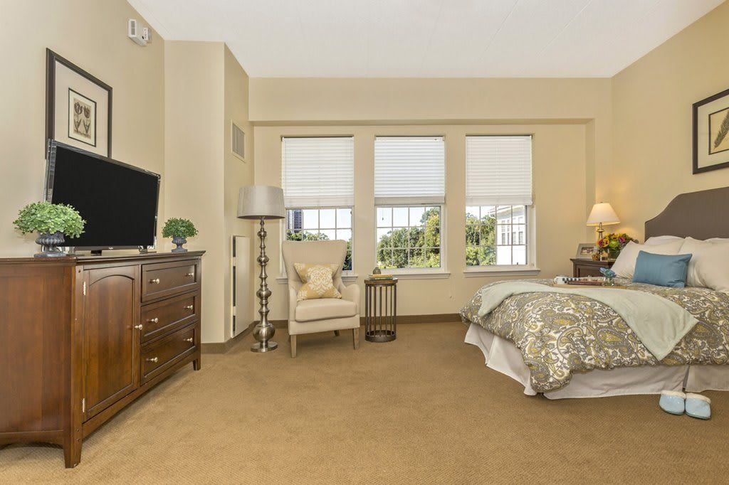 A cozy and elegantly designed bedroom with a bed, armchair, television, and large windows.